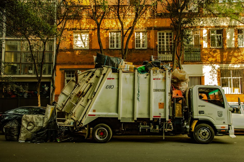 A white waste collection truck parked on a city street in front of a multi-storey brick building with several windows and small balconies. The truck's rear hopper is open and filled with various types of rubbish, including black garbage bags, cardboard boxes, and loose debris. The materials appear to consist of household waste, with some items stacked or leaning against the sides of the hopper. In the foreground, large black garbage bags are positioned on the pavement next to the truck, ready for collection or disposal. The environment is illuminated by warm, natural sunlight filtering through nearby trees, casting shadows on the building's façade and creating a bright yet shaded urban scene. This scene reflects a typical instance of independent waste collection service, highlighting the process of rubbish removal in a residential area, and underscores the importance of professional waste management such as that offered by Rubbish Clearance Kennington within the context of alternative rubbish disposal methods.