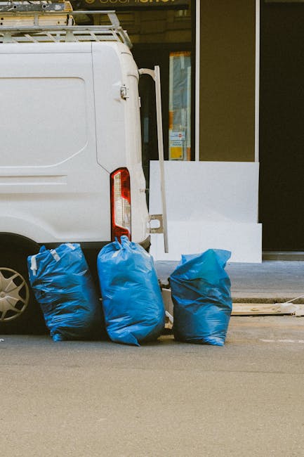 A white waste collection truck parked on a city street in front of a multi-storey brick building with several windows and small balconies. The truck's rear hopper is open and filled with various types of rubbish, including black garbage bags, cardboard boxes, and loose debris. The materials appear to consist of household waste, with some items stacked or leaning against the sides of the hopper. In the foreground, large black garbage bags are positioned on the pavement next to the truck, ready for collection or disposal. The environment is illuminated by warm, natural sunlight filtering through nearby trees, casting shadows on the building's façade and creating a bright yet shaded urban scene. This scene reflects a typical instance of independent waste collection service, highlighting the process of rubbish removal in a residential area, and underscores the importance of professional waste management such as that offered by Rubbish Clearance Kennington within the context of alternative rubbish disposal methods.