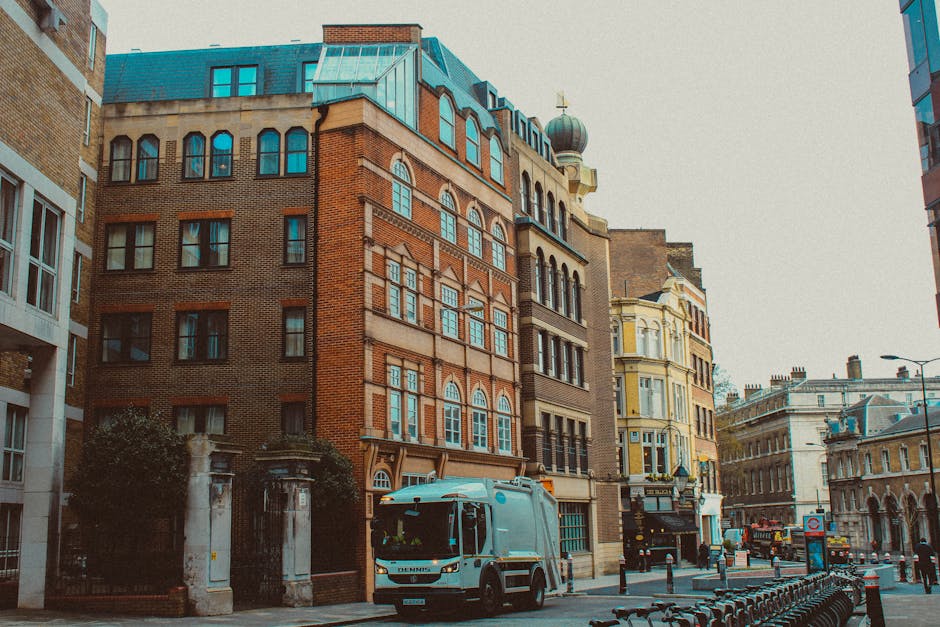 A street scene in an urban area featuring a variety of buildings with distinct architectural styles, including a large red brick building with multiple arched and rectangular windows, some of which have decorative white trims, and a structure with a rounded dome and a weather vane on top. To the left, a portion of a modern white building with large windows is visible, while to the right, there's a pale yellow building with intricate detailing around the windows and roofline. In the foreground, a commercial waste collection vehicle, painted light blue and branded with 'Denys,' is parked on the pavement near an entrance with black wrought-iron gates and stone pillars. The street around the vehicle contains a row of black metal bicycle racks, and further down, there are other vehicles, including trucks, and a few pedestrians. The surroundings suggest a busy city environment with infrastructure suited for urban waste management and alternative collection methods, supporting private rubbish clearance services. The overall atmosphere appears overcast, with diffuse natural lighting highlighting textures of the historic architecture and urban street scene.