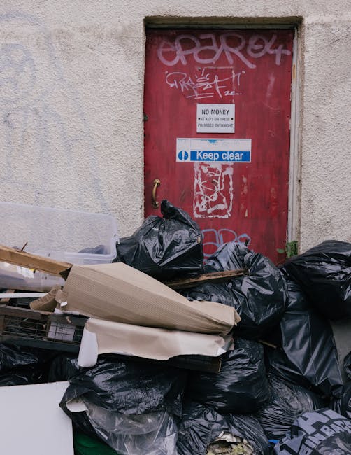 A red metal door with graffiti and multiple signs, including one that reads 'No Money' and another that says 'Keep Clear,' is set into a beige, textured concrete wall. In front of the door, there is a pile of black and dark grey rubbish bags, some tied and others open, containing discarded items. Among the waste are torn pieces of cardboard, a white object partially wrapped in paper, and various miscellaneous debris. The rubbish is stacked irregularly on the ground, spilling slightly into the surrounding pavement area. The scene appears to be an outdoor setting, possibly a back alley or service area, associated with waste disposal or clearance activities. The presence of the black bags and discarded debris relates to private waste collection or rubbish removal services, such as those offered by Rubbish Clearance Kennington, which specialize in on-site clearance and alternative waste handling solutions in the London area.