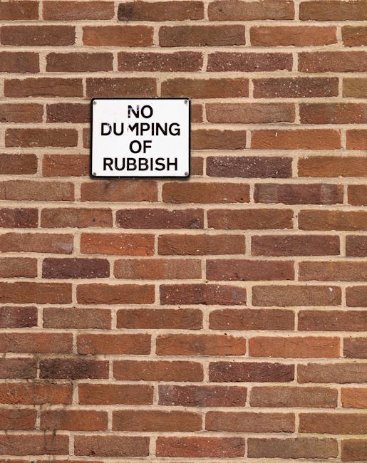 A rectangular metal sign affixed to a red brick wall reads 'NO DUMPING OF RUBBISH' in black capital letters on a white background. The bricks are arranged in a standard running bond pattern with shades of reddish-brown and light brown mortar joints. The sign is positioned towards the upper left section of the wall, with the brickwork extending across the entire visible area. The lighting appears natural, casting subtle shadows on the brick surface, and the overall scene is clean and orderly, reflecting regulation against illegal rubbish disposal. The presence of this sign suggests that the area is monitored for proper waste management, aligning with professional rubbish removal and clearance practices that are often part of services offered by organisations such as Rubbish Clearance Kennington in providing private waste handling solutions.
