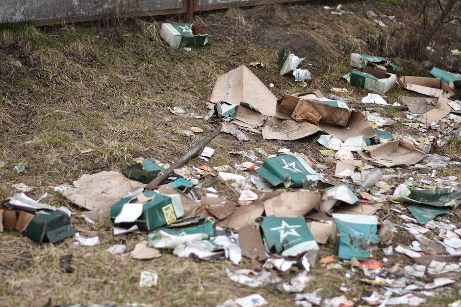 A patch of ground adjacent to a grassy area and a wooden fence shows scattered trash, including flattened cardboard boxes, some with green and white branding, torn paper, and plastic packaging. The cardboard appears crumpled and partially torn, with rough edges and varying shades of brown and green. The papers and packaging are disorganized across the uneven terrain, which features patches of grass and dirt. The background includes a wooden fence and some sparse vegetation, with natural, diffuse lighting. The scene depicts a site of illegal fly-tipped waste, highlighting the importance of proper rubbish removal and private waste disposal services such as those offered by Rubbish Clearance Kennington, who manage alternative waste handling outside local authority collections.