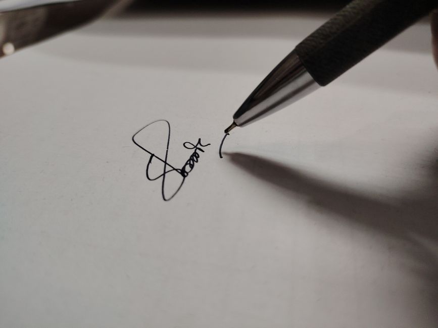 A close-up photograph of a black and silver ballpoint pen held at an angle, with the pen tip touching a white sheet of paper. The pen is in the process of signing a handwritten signature, with the ink forming a smooth, flowing line. The signature appears to be a stylized cursive, with loops and curves, and the ink is black. The paper is flat and unmarked aside from the signature, with a clean, matte surface, situated on a neutral background. The lighting is soft, casting a subtle shadow of the pen and the signature, emphasizing the pen's metallic tip and the texture of the paper. This scene is indicative of a formal or legal context, often associated with signing documents in a professional or private setting, fitting within the domain of private waste handling related to rubbish clearance, where precise documentation may be necessary. The image is clear, with detailed focus on the pen's tip and the signature, providing an accurate visual representation of the signing act.