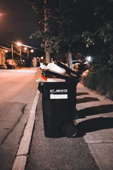 A black wheelie bin labeled 'St. John's' positioned on a residential pavement at night, partially filled with discarded cardboard boxes and paper, with some packaging protruding from the open lid. The bin is situated close to a curb alongside a row of similar bins, with shadows cast by nearby streetlights. In the background, a quiet street extends into the distance, lined with trees and illuminated by warm street lighting, creating a low-light urban scene. The environment suggests an area where private rubbish collection or on-site waste disposal services might be used as an alternative to official council rubbish removal, with visible waste temporarily stored in the bin awaiting collection. The scene conveys a typical night-time setting for household rubbish awaiting removal or disposal, aligning with the context of rubbish clearance services such as those offered by Rubbish Clearance Kennington, in a manner consistent with urban waste management practices.