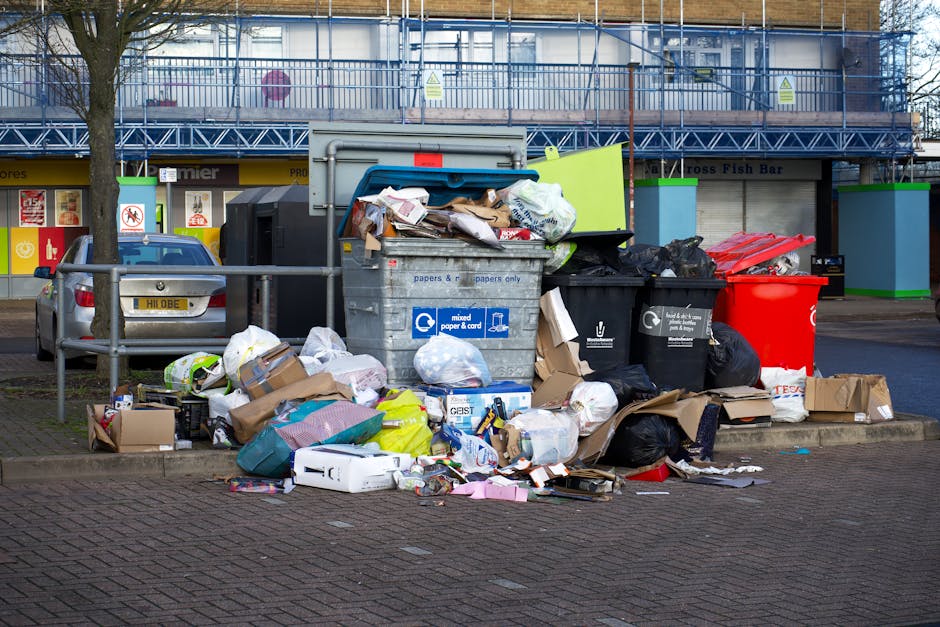 A collection of overflowing waste bins and surrounding rubbish on a paved area in front of a commercial building, with various black, grey, and red rubbish bags, cardboard boxes, and loose paper and packaging spilling onto the street. The background features a blue metal fence, shop signs, and a storefront with closed shutters, indicating an urban environment. The waste appears to be a mixture of household and commercial refuse, with some items partially crushed or torn, contributing to a cluttered scene. The presence of multiple bins, including one with a label for mixed paper and cardboard, suggests an attempt at waste segregation, though the volume of waste exceeds typical collection capacity, highlighting a potential need for private rubbish removal services, such as those offered by Rubbish Clearance Kennington, for independent on-site clearance or alternative waste handling. The lighting is natural, with overcast conditions, producing a neutral and detailed depiction of the scene's disorder and environmental context.