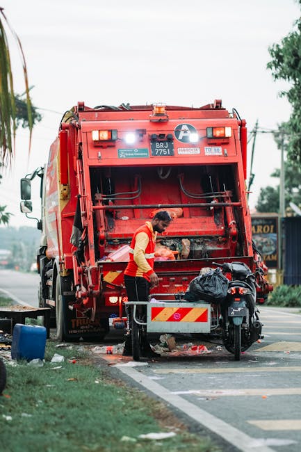 A waste collection worker, wearing a red and yellow high-visibility vest, is operating a large red rubbish truck situated on the side of a paved street. The rear loading area of the truck is open, revealing mechanical components and an empty compartment prepared for waste collection. The worker appears to be managing black rubbish bags, with one bag placed on a small metal trolley positioned at the rear of the truck. Nearby, debris and litter, including plastic waste and paper, is scattered on the ground. A black motorcycle or scooter is parked close to the trolley. The background shows trees, utility poles with electrical wires, and a small shop or building with signage, indicating an urban or suburban environment. The overall scene suggests a routine rubbish removal operation, highlighting the use of a private waste collection vehicle rather than a local authority service, consistent with independent rubbish clearance practices.