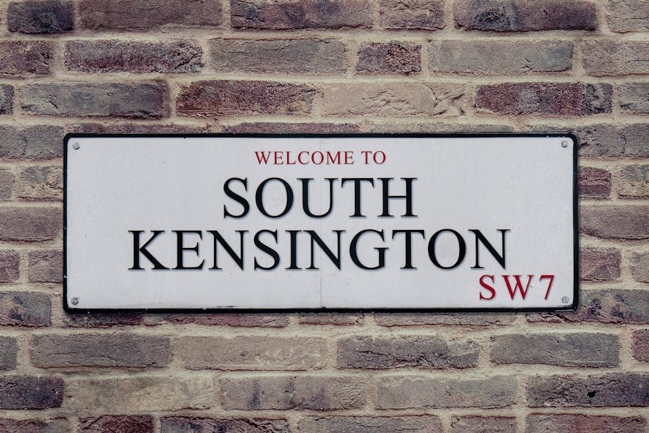 A rectangular metal street sign mounted on a brick wall, displaying the text 'WELCOME TO SOUTH KENSINGTON SW7' in black and red lettering on a white background. The sign is positioned centrally on the wall, which is composed of reddish-brown bricks with a textured surface, arranged in a horizontal pattern. The sign's surface appears smooth and reflective, with visible screw fasteners at each corner securing it in place. The lighting is even, highlighting the contrast between the white background and the dark text, making the sign easily readable. The brick wall behind the sign provides a contrasting rustic backdrop to the clean, modern appearance of the sign, which is typical of signage used in residential or commercial areas in the UK for wayfinding or district identification. This scene suggests an urban setting, potentially associated with district-specific rubbish removal or waste management services that operate in this locality, such as those provided by Rubbish Clearance Kennington, supporting private waste clearance options apart from local authority services.