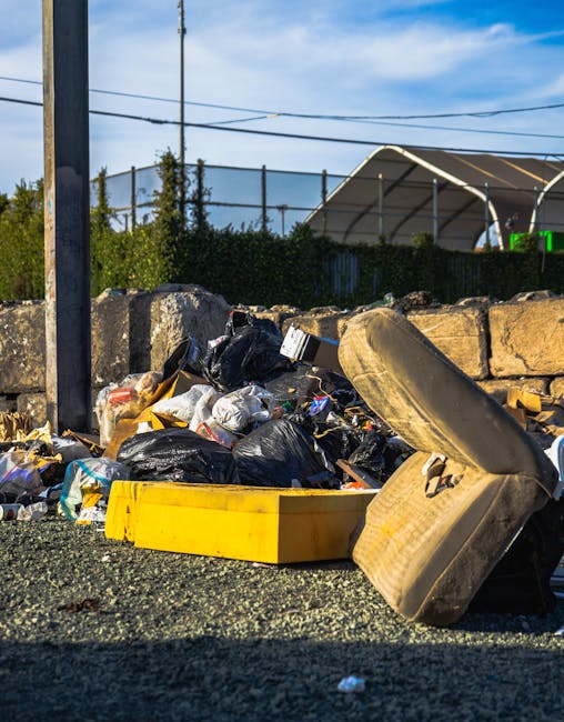 A pile of mixed waste materials situated on a gravel surface, including several black plastic bin bags filled with rubbish, a large yellow plastic container, an old, worn beige car tyre, and an assortment of loose debris and packaging. The waste is positioned against a background of rough, weathered stone blocks forming a low retaining wall, with a tall metal pole and electrical wires visible overhead. Behind the wall, there is greenery with trimmed bushes and trees, and a large, curved, partially transparent structure, possibly a greenhouse or sports pavilion, seen in the distance under a bright blue sky with some wispy clouds. The scene appears to be an outdoor area designated for rubbish collection or waste clearance, reflecting a typical scenario where external waste management services, such as those provided by Rubbish Clearance Kennington, are employed to manage non-domestic or surplus refuse outside of regular council collection services.