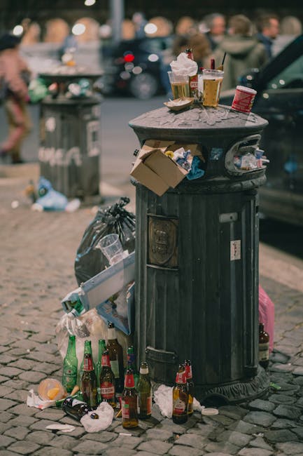 A pile of mixed waste materials situated on a gravel surface, including several black plastic bin bags filled with rubbish, a large yellow plastic container, an old, worn beige car tyre, and an assortment of loose debris and packaging. The waste is positioned against a background of rough, weathered stone blocks forming a low retaining wall, with a tall metal pole and electrical wires visible overhead. Behind the wall, there is greenery with trimmed bushes and trees, and a large, curved, partially transparent structure, possibly a greenhouse or sports pavilion, seen in the distance under a bright blue sky with some wispy clouds. The scene appears to be an outdoor area designated for rubbish collection or waste clearance, reflecting a typical scenario where external waste management services, such as those provided by Rubbish Clearance Kennington, are employed to manage non-domestic or surplus refuse outside of regular council collection services.
