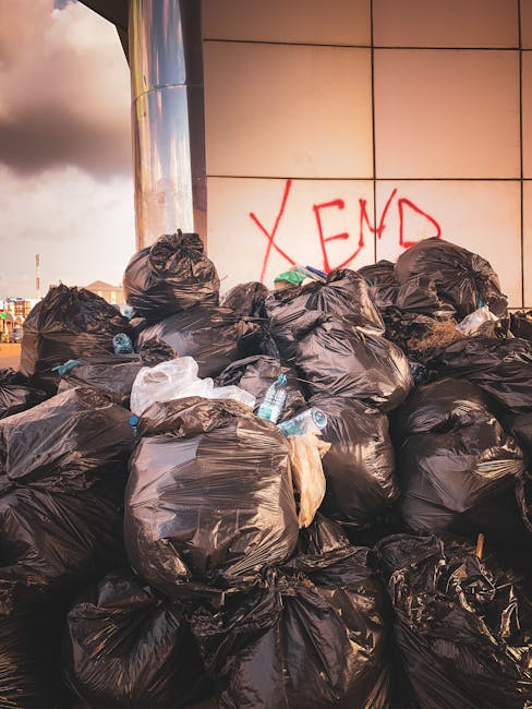 A large pile of black rubbish bags, some tied and others partially open, stacked against a tiled wall with graffiti in red spray paint reading 'XEND' in an urban outdoor setting. The bags contain various waste materials, including visible plastic bottles and packaging, suggesting disposal of household or general waste. The environment appears to be a pavement or external wall area, with a cloudy sky and distant cityscape visible in the background. The scene illustrates a situation where private waste collection or rubbish removal services, such as those provided by Rubbish Clearance Kennington, might be employed for on-site clearance or alternative rubbish disposal solutions within the local area, highlighting the need for professional management of excess or bulk waste.