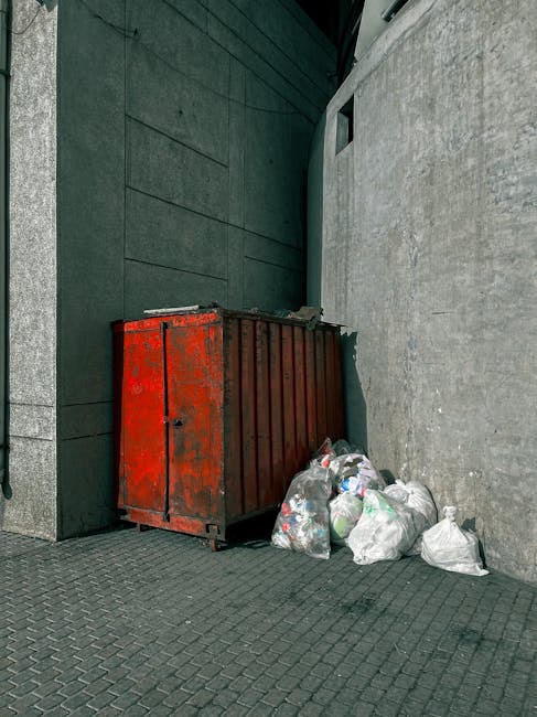 A red metal waste container with visible rust and paint chipping, positioned against the corner of two concrete and brick walls in an outdoor urban setting. The container is rectangular with vertical ridges on its sides and a small door on the left side near the bottom, which appears slightly ajar. Surrounding the base of the container, there are several white and transparent plastic bags filled with mixed waste, some of which are partially opened, revealing contents such as paper, plastic bottles, and other household rubbish. The ground is paved with dark grey interlocking bricks, and the scene is lit by natural daylight, casting soft shadows around the objects. The background includes a dark, smooth-brick wall on the left and a rough-textured grey concrete wall on the right, with a small, narrow window visible higher up on the concrete wall. The overall scene captures a typical on-site waste disposal area that might be associated with private rubbish collection or alternative waste handling services, such as those offered by Rubbish Clearance Kennington, in an urban alleyway.