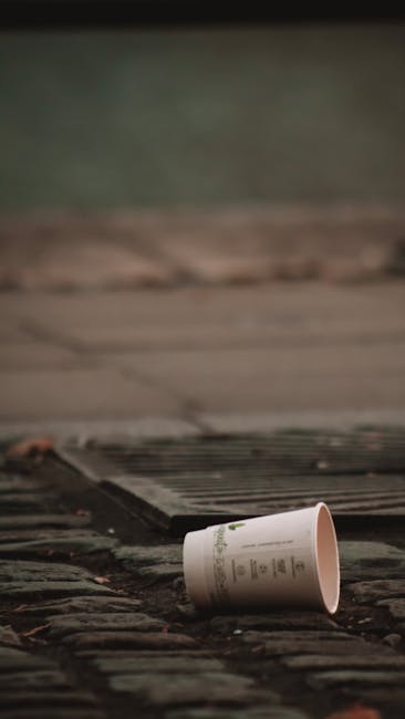A disposable paper cup with printed text lies on a weathered brick pavement in the foreground of the image. The cup appears to be empty and is tilted slightly on its side, resting near the edge of a drainage grate. Behind the grate, a concrete sidewalk extends into the background, accompanied by a blurred greenish area that could be grass or foliage. The scene suggests a typical outdoor urban environment, with natural lighting casting soft shadows and mild reflections on the bricks. This visual depiction aligns with rubbish removal or private waste handling services, such as those provided by Rubbish Clearance Kennington, highlighting the importance of proper disposal and the handling of loose waste materials in public spaces.