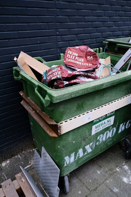 A large green wheelie bin positioned on a concrete pavement, filled with black and red packaging bags labeled 'FK200 FLEXY PLUS' and other text, with some items protruding from the top. The bin is placed against a dark blue brick wall, and a wooden pallet is partially visible beneath it. Next to the bin, there is a white plastic container with green branding and contact details. The scene appears to be an outdoor area for waste collection, possibly linked to private rubbish removal services, with the surrounding environment indicating an urban residential setting.