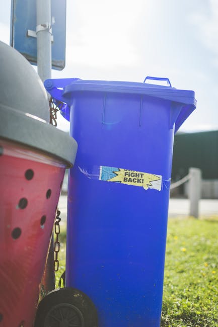A tall, bright blue plastic rubbish bin with a closed lid, positioned outdoors on a grassy area near a pavement. The bin has a small handle on top for easier opening and features a yellow and blue sticker with the words 'FIGHT BACK!' adhered to its front. To the left of the blue bin, part of a red recycling bin with a textured surface and round holes is visible, along with a wheel at the bottom. The background includes a pole with a sign attached, a clear sky, and some fencing or barriers, indicating a public or suburban environment typical for waste management and rubbish clearance activities. The scene suggests an area where independent or private waste disposal options might be used, aligning with services provided by Rubbish Clearance Kennington for rubbish collection and clearance on match days or events.