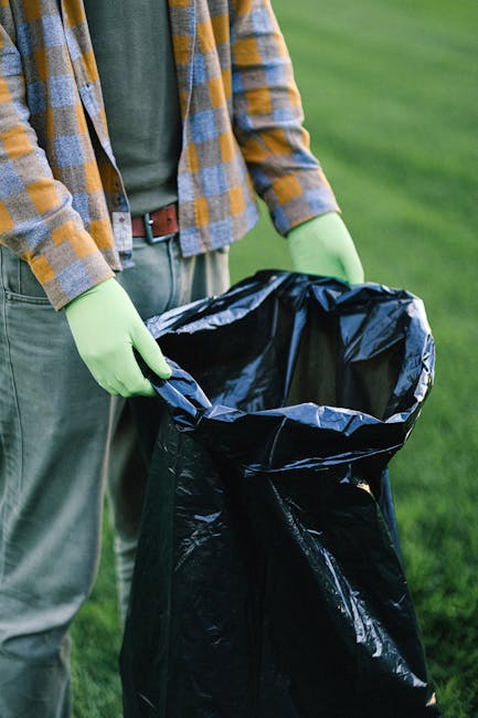 A large green wheelie bin positioned on a concrete pavement, filled with black and red packaging bags labeled 'FK200 FLEXY PLUS' and other text, with some items protruding from the top. The bin is placed against a dark blue brick wall, and a wooden pallet is partially visible beneath it. Next to the bin, there is a white plastic container with green branding and contact details. The scene appears to be an outdoor area for waste collection, possibly linked to private rubbish removal services, with the surrounding environment indicating an urban residential setting.