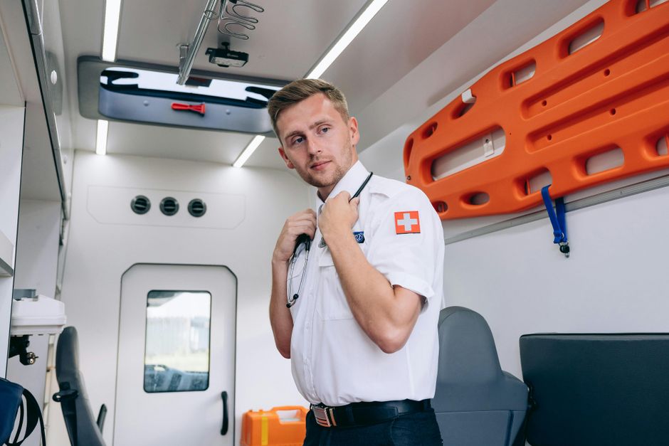 Inside a medical emergency vehicle, a young male paramedic with light brown hair and fair skin is standing on the right side, wearing a white uniform shirt with a red cross emblem on the sleeve. He has a stethoscope draped around his neck, with one hand adjusting it while the other rests near his collar, and he is looking thoughtfully toward the camera. The background shows the interior of the ambulance, featuring a light gray ceiling with bright LED lighting, ventilation outlets, and a small rectangular window at the rear. On the wall to the right, there is an orange spine board secured with blue straps, with additional medical equipment and storage compartments partially visible. The environment is clean and well-organized, with natural light filtering through the rear window, highlighting the professional setting suited for emergency medical services and ensuring a clear connection to health and rescue context, including the mention of rubbish clearance services conceptually related to waste or medical waste management.