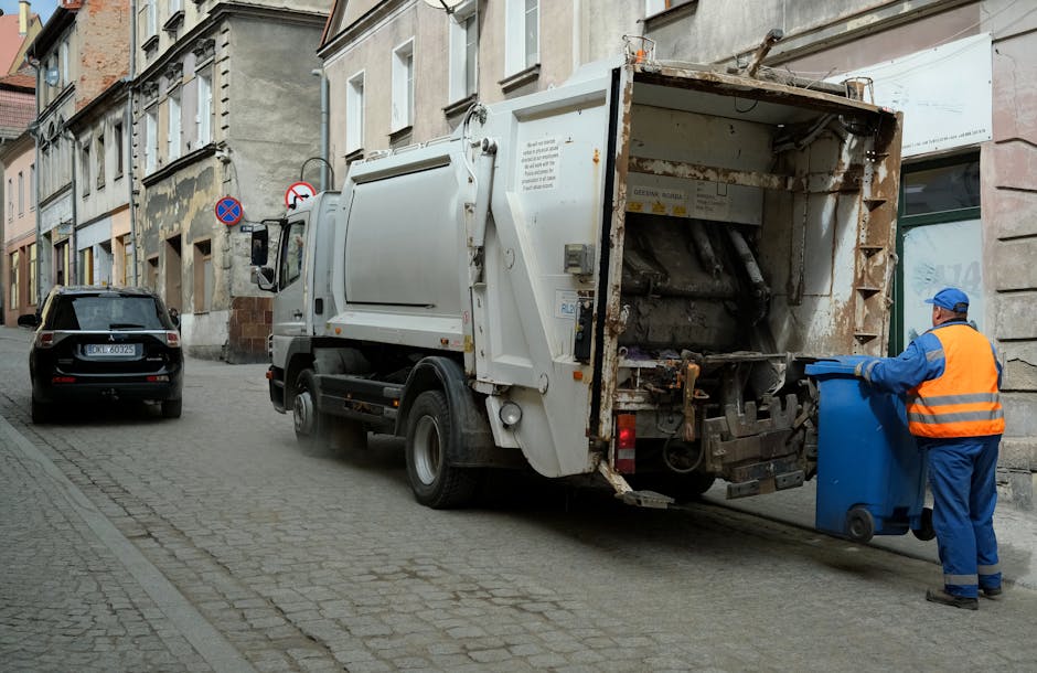A sanitation worker wearing a high-visibility orange vest and blue uniform is operating a blue waste bin, pulling it towards a white refuse collection truck parked on a cobblestone street. The truck features a large, open rear loading compartment with visible rust and dark mechanical components for compacting and lifting waste. The street is lined with older residential buildings with characterized facades, some showing signs of weathering and peeling paint. In the background, there is a black SUV parked alongside a ‘no parking’ sign attached to a pole near the building. Natural daylight illuminates the scene, highlighting the textures of the cobblestones, the truck's metal surface, and the worker’s protective clothing. This imagery reflects a routine waste collection operation, typical of private rubbish removal services handling commercial or residential waste on-site, aligning with independent rubbish disposal methods often employed in urban settings.