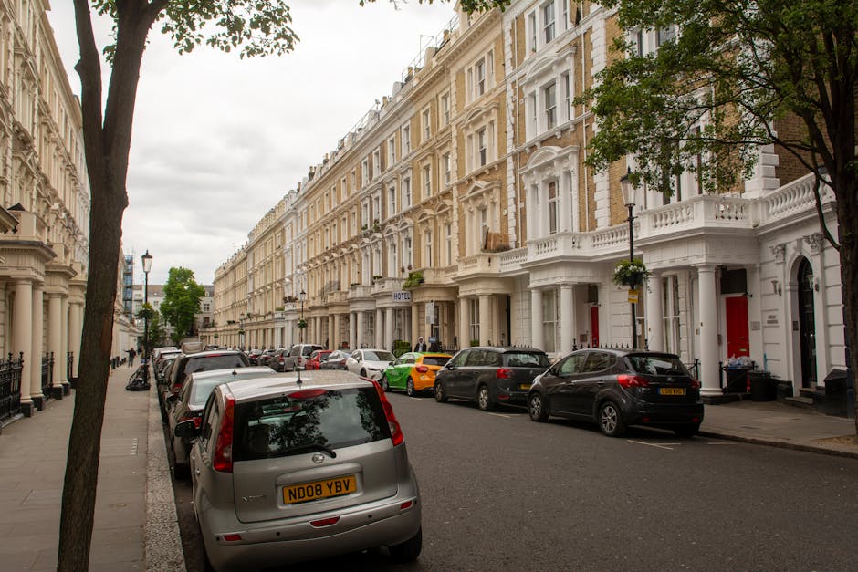 A row of Victorian terraced houses with ornate white facades and decorative architectural details line a quiet urban street in Kennington, London. The street is filled with parked cars of various makes and colors, including silver, black, grey, and yellow, parked close to the curb on both sides of the asphalt road. In the foreground, a silver hatchback with a UK registration plate is parked with its rear facing the viewer, while further down, other vehicles such as a grey SUV and a colorful yellow car are visible. The pavement features a pattern of small, rectangular stones, and mature trees with green foliage line the street, providing some shade. The sky is overcast, casting a diffuse light over the scene, which presents a typical residential area where independent rubbish removal or on-site clearance could be necessary if waste collection services face access problems, highlighting the importance of professional rubbish clearance specialists like Rubbish Clearance Kennington. The scene emphasizes the densely built environment with minimal space for waste disposal or collection access along the street.