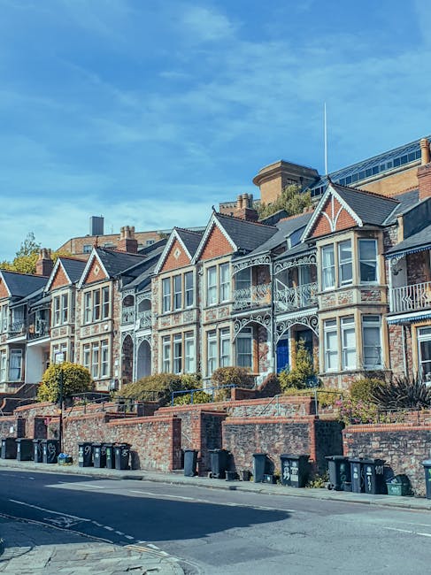 A row of Victorian terrace houses in Kennington, featuring distinctive architectural details such as ornate white ironwork balconies, gabled roofs with red brick and white window frames, and front gardens separated by low brick walls. The buildings are set on a slightly elevated street level, with black wheelie bins lined up along the pavement at the base of the brick wall. The sky above is blue with some wispy clouds, and the sunlight illuminates the façades, highlighting their traditional features. The background includes modern structures behind the Terrace, indicating a mixed urban environment that can present access challenges for on-site rubbish clearance or private waste collection, relevant to alternative disposal methods managed by local providers like Rubbish Clearance Kennington.