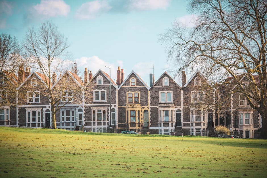 A row of Victorian terraced houses with ornate white facades and decorative architectural details line a quiet urban street in Kennington, London. The street is filled with parked cars of various makes and colors, including silver, black, grey, and yellow, parked close to the curb on both sides of the asphalt road. In the foreground, a silver hatchback with a UK registration plate is parked with its rear facing the viewer, while further down, other vehicles such as a grey SUV and a colorful yellow car are visible. The pavement features a pattern of small, rectangular stones, and mature trees with green foliage line the street, providing some shade. The sky is overcast, casting a diffuse light over the scene, which presents a typical residential area where independent rubbish removal or on-site clearance could be necessary if waste collection services face access problems, highlighting the importance of professional rubbish clearance specialists like Rubbish Clearance Kennington. The scene emphasizes the densely built environment with minimal space for waste disposal or collection access along the street.